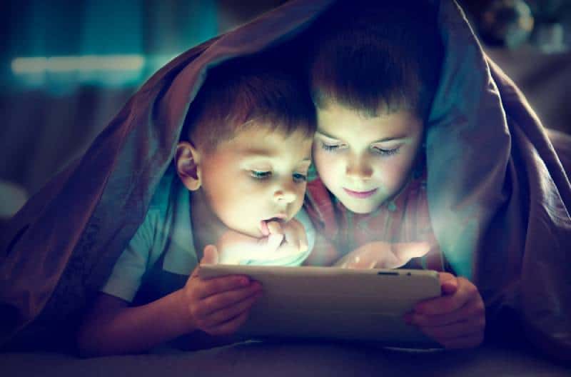 Two kids using tablet pc under blanket at night