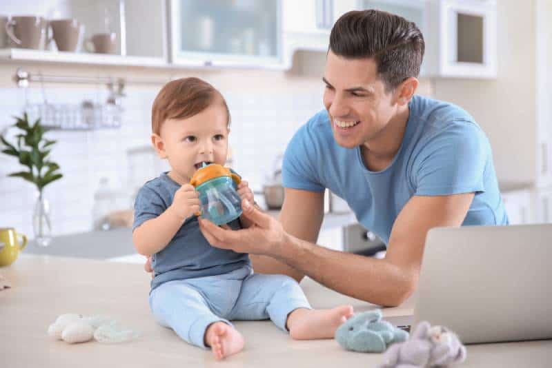 Young dad giving water in a sippy cup to his little son at home
