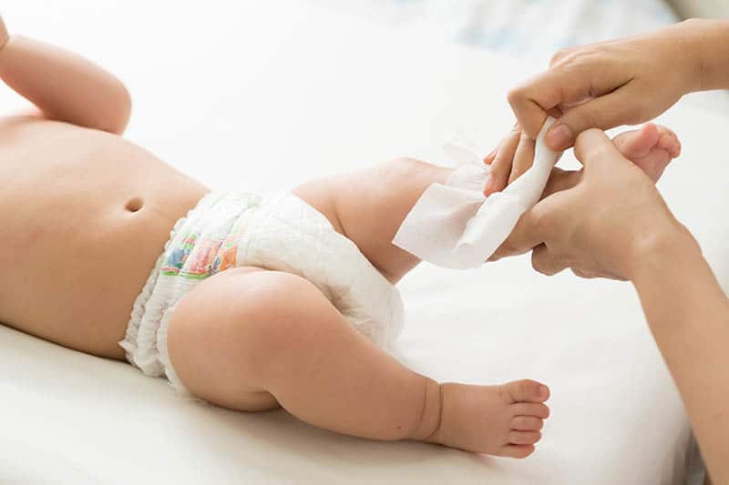 Mom is wiping baby's leg while he is lying on bed in a diaper only