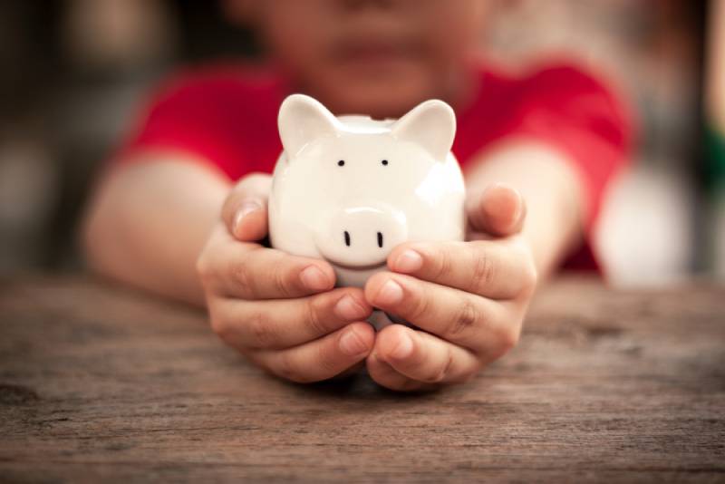 Child's hand is holding a white piggy bank on wooden table