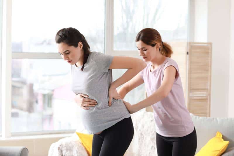 Doula giving a back massage to a pregnant woman in pain in her home next to the white sofa with yellow pillows