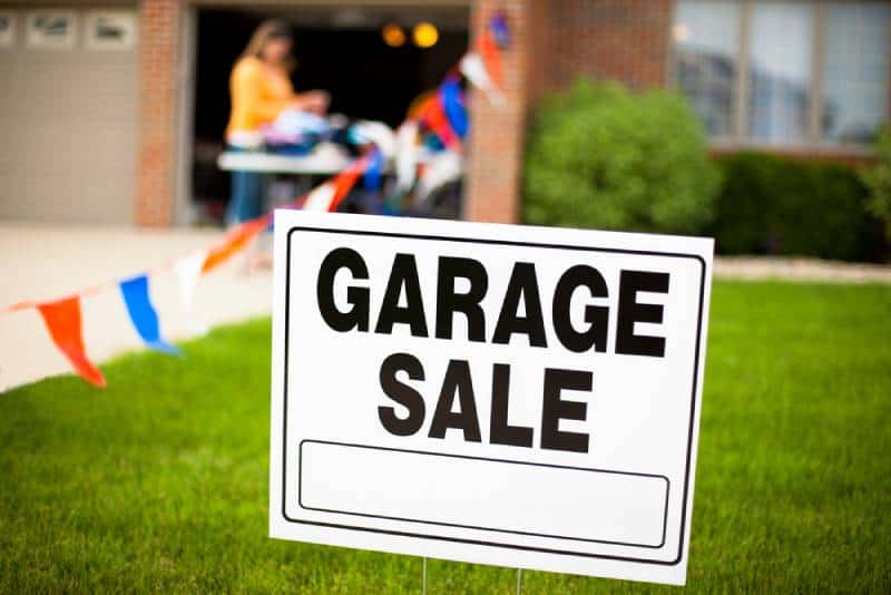 Garage sale sign on the front yard of a suburban house with a woman looking at items on a table