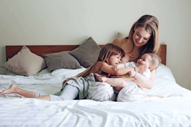 Happy mom playing on the bed and hugging her 2 kids, enjoying together