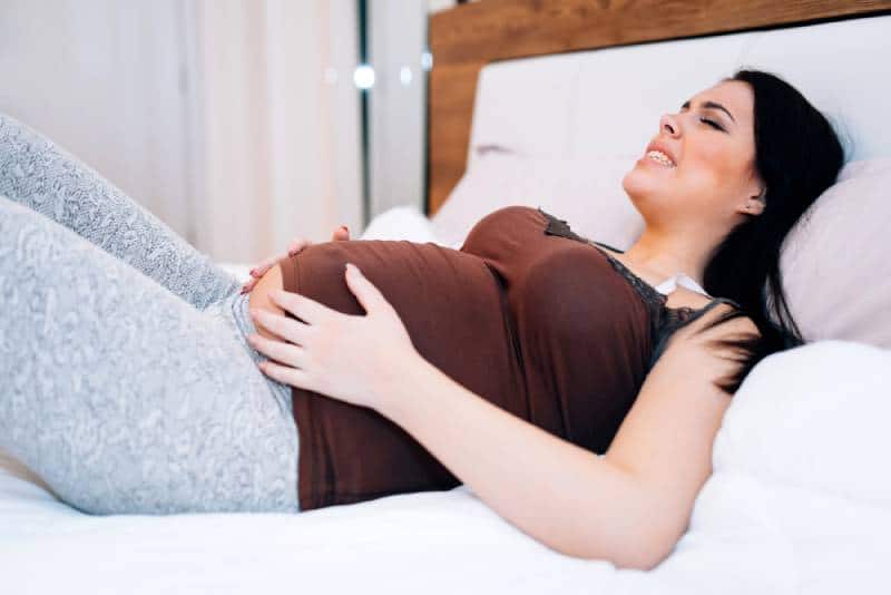 Pregnant woman having cramps and holding her tummy while lying on bed at home