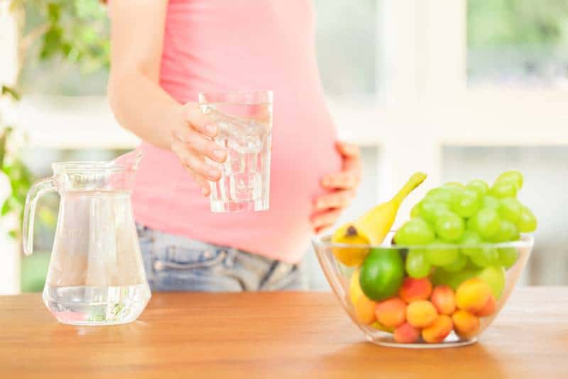 Pregnant woman with a glass of water and bowl of fruits in kitchen