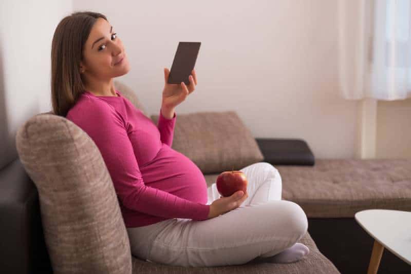 Pregnant woman sitting on sofa in pink shirt holding chocolate and an apple