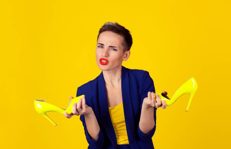 Confused woman holding pair of high heels shoes on a yellow background