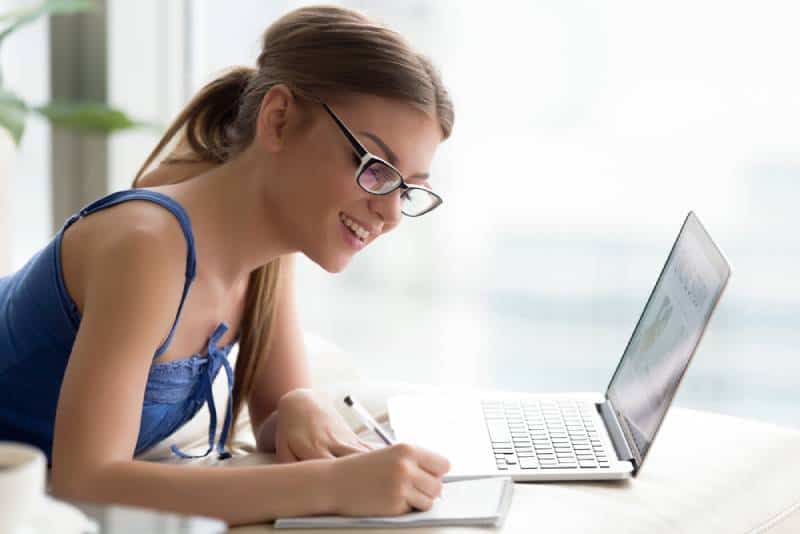 young woman wearing glasses studying online on laptop, writing down in notebook important information from course