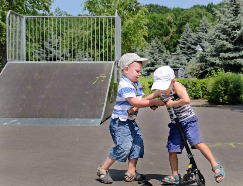 Two young boys in baseball caps fighting over a scooter in a skate park