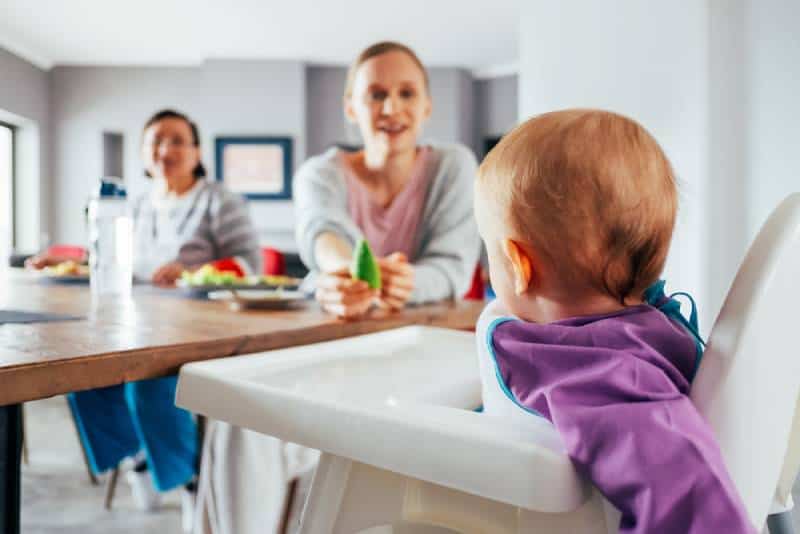 Young mom feeding her child with solid food in dining room while cute baby girl sitting on highchair and having breakfast