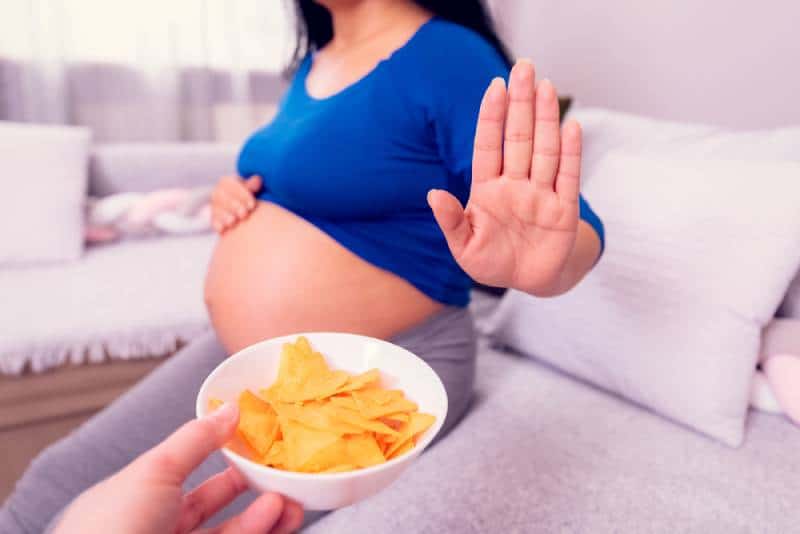 Pregnant woman refuses to eat unhealthy chips while sitting on a bed in her room