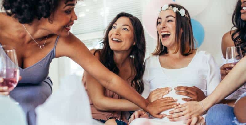 Laughing pregnant woman sitting with friends touching her tummy at gender reveal games party