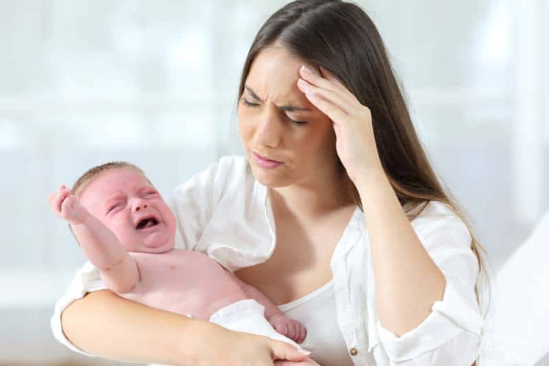 Sick mom in white holding crying baby while having a severe headache