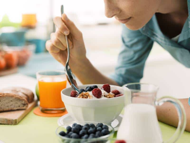 Woman having an healthy delicious breakfast at home, she is eating yogurt with cereals and fresh fruit, healthy food concept