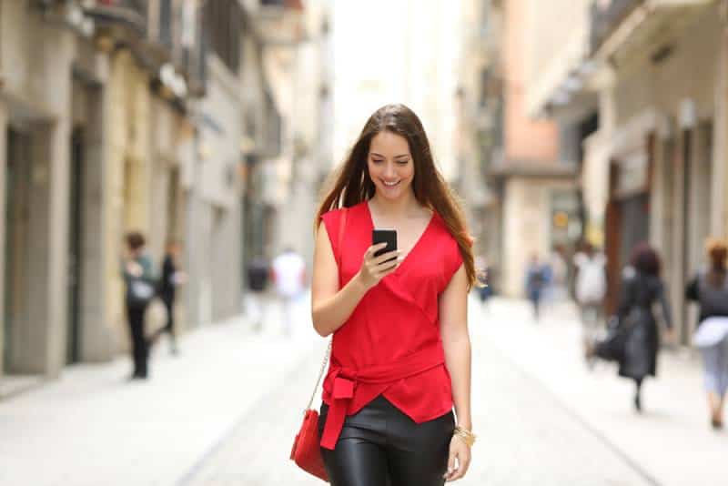 Front view of a fashion happy woman in red shirt and black pants walking and using a smart phone on a city street