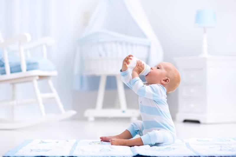 baby boy playing on a blue floor mat and drinking milk from a bottle in a white sunny nursery with rocking chair and bassinet