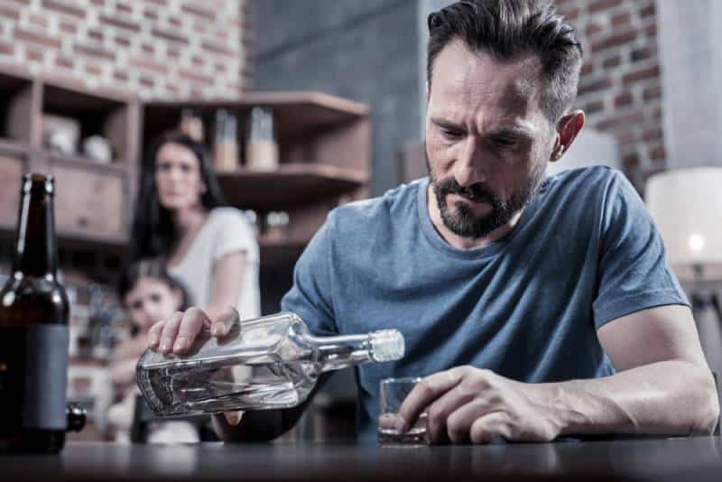 Serious unhappy sad man sitting at the table and pouring vodka into his glass while his wife and daughter watch