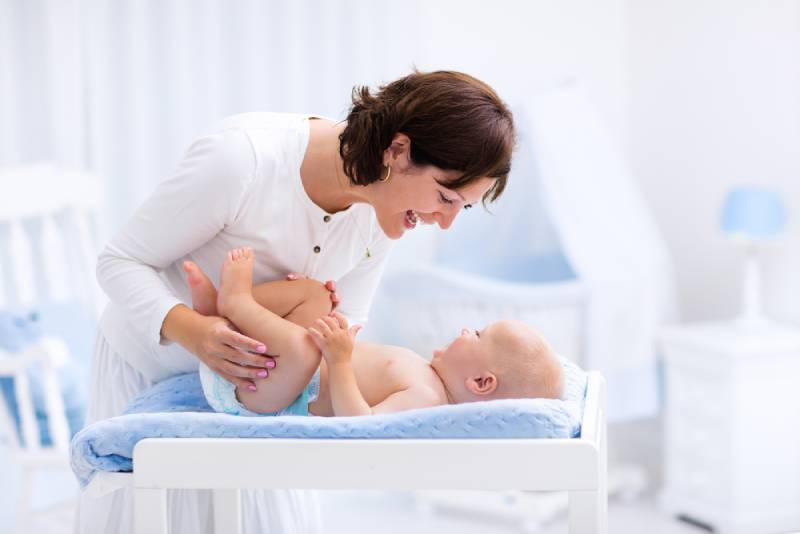 mother taking care of little boy in white sunny nursery with changing table, baby crib and rocking chair
