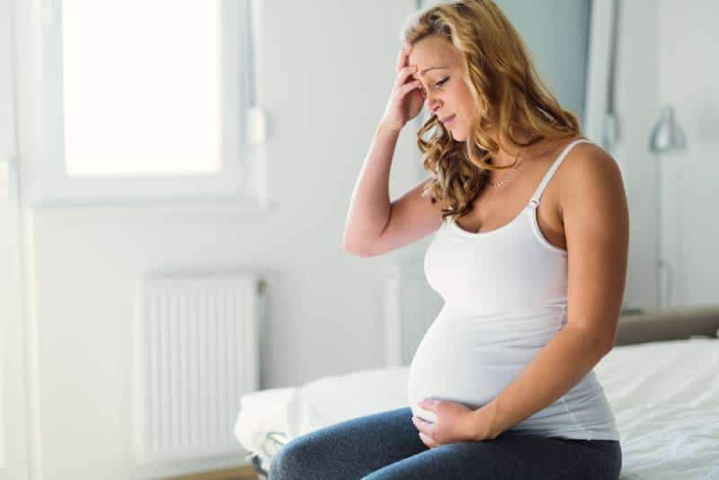 Pregnant woman in jeans and white shirt suffering with headache and nausea while sitting on bed in her room