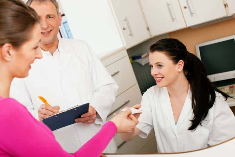 Patient in reception area of office of doctor or dentist, handing her health insurance card over the counter to the nurse, while doctor is there