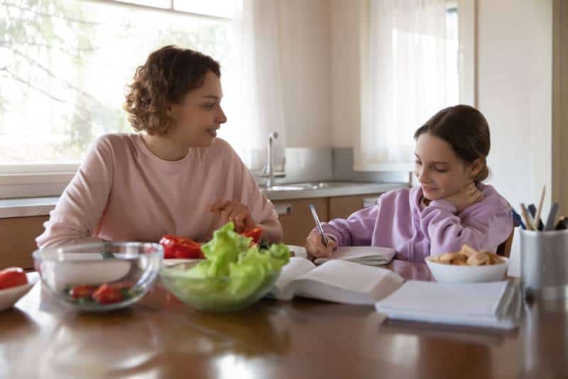mum preparing vegetable salad helping tween school girl child doing homework sitting at kitchen table