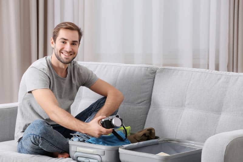 Man in grey shirt and denim sitting on a sofa and packing his suitcase