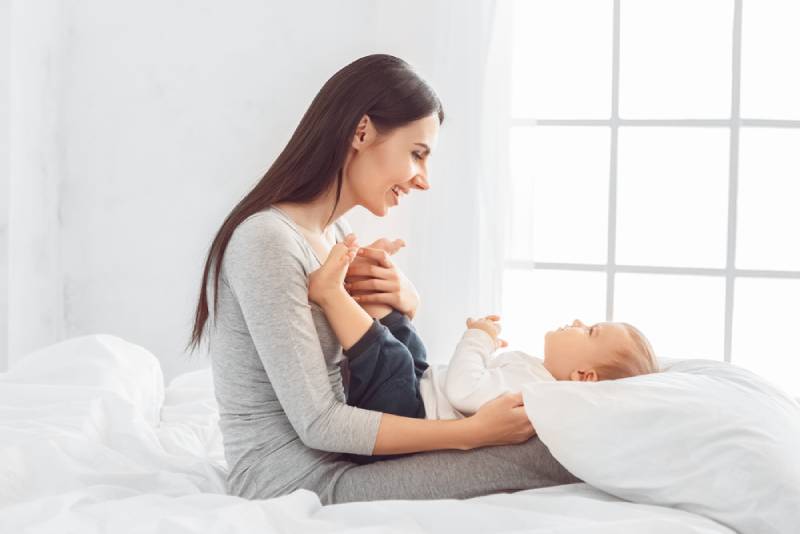 Mother sitting with son lying on her knees on bed smiling joyful in a room