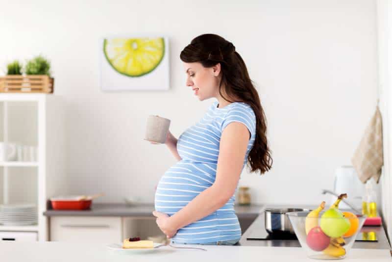 pregnant woman with cup of tea and piece of cake at home kitchen