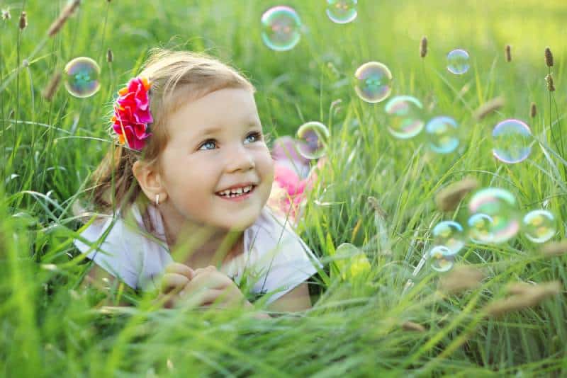 three year old girl laying on a grass in a park playing with bubbles