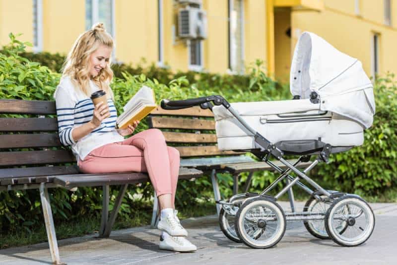mother reading book and holding coffee to go on bench near baby stroller in park