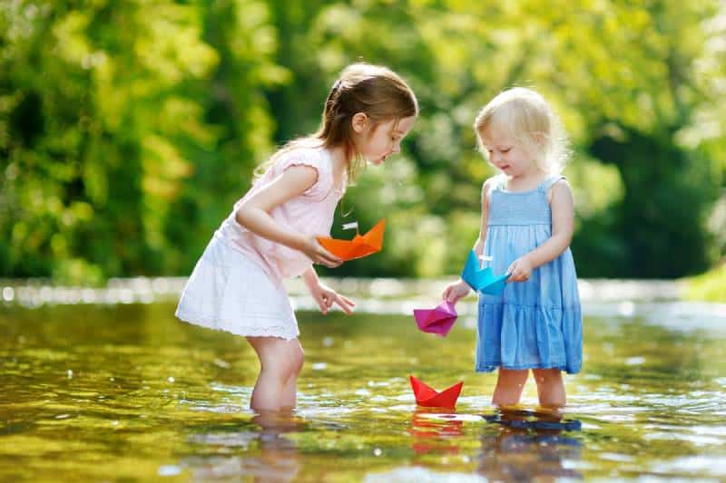 Two adorable little sisters playing with paper boats by a river 