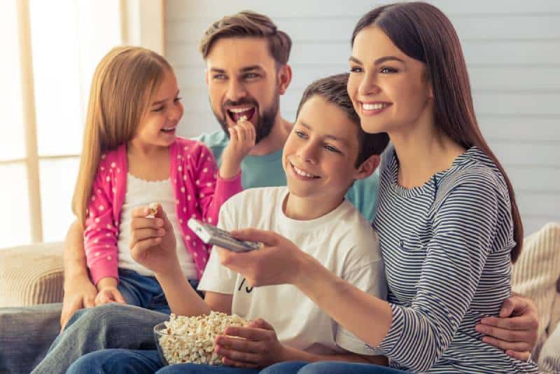 young parents, their daughter and son are watching TV, eating popcorn and smiling