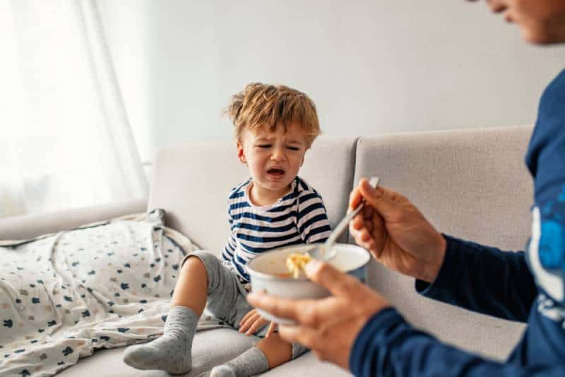  child refusing to eat at home with white background while father is trying to deceive the toddler by tricks