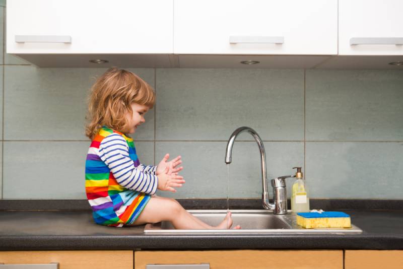  toddler girl in a colorful dress washes her hands and playing with foam in the sink in a kitchen