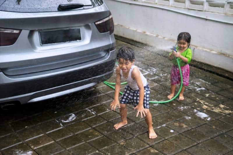 Little girl playing a water hose while washing a car with her brother at home