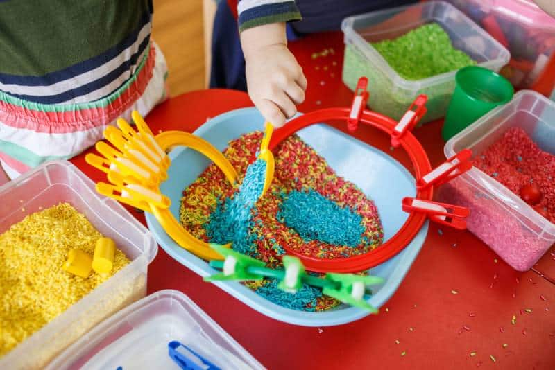 Toddlers playing with sensory bin with colourful rice on red table