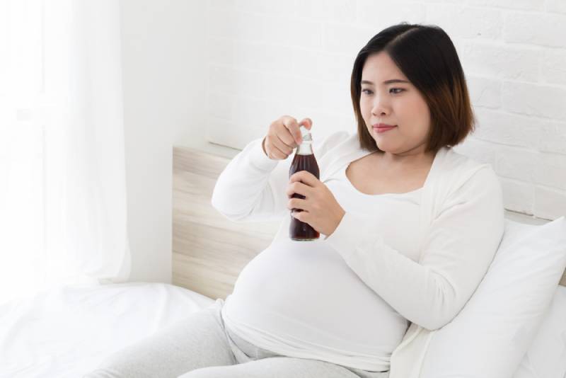 Pregnant woman in white outfit drinking cola and lying on bed