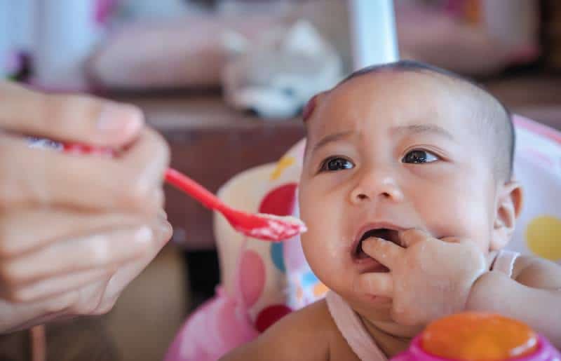 baby refusing to eat baby food with a red spoon on a baby chair and crying
