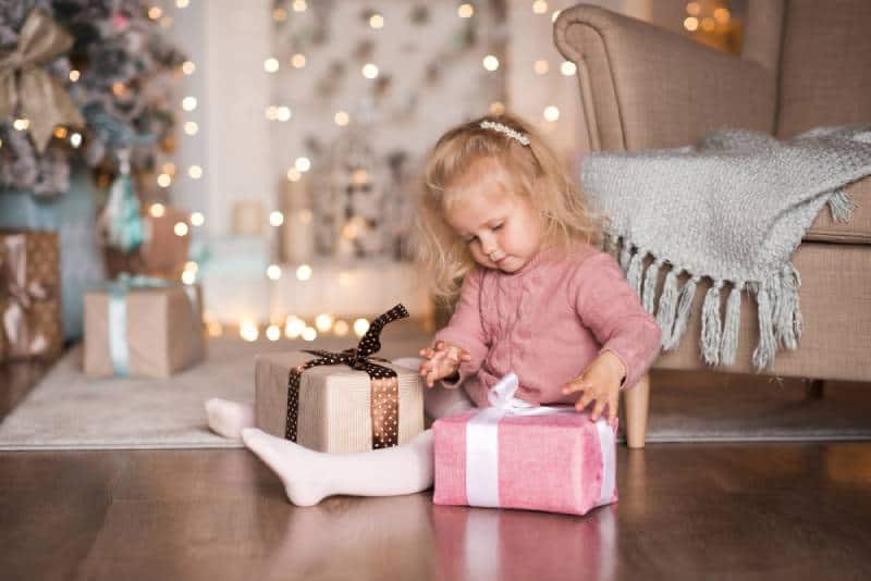 3 year old girl open Christmas present sitting on floor in room over Christmas decor