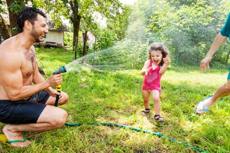Dad and mom play with a little daughter outdoors in the summer, pouring water from a hose