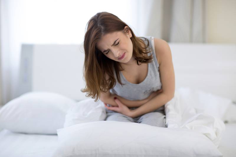 Young woman sitting on the bed and holding her stomach with pain