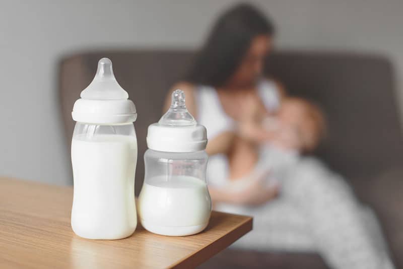 two baby bottles on the table while woman feeding baby behind
