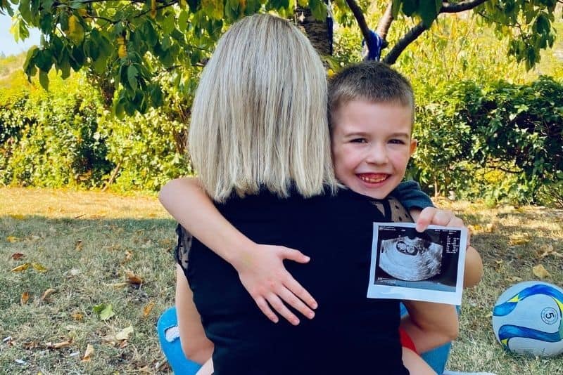 Boy sitting in mom's lap with sonogram of his brother or sister outdoors