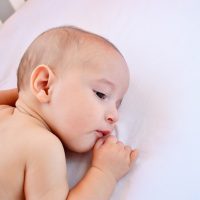 cute baby boy lying on white sheet in crib and sucking lip