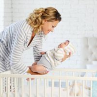 Mom puts her baby to crib and smiling in a nursery