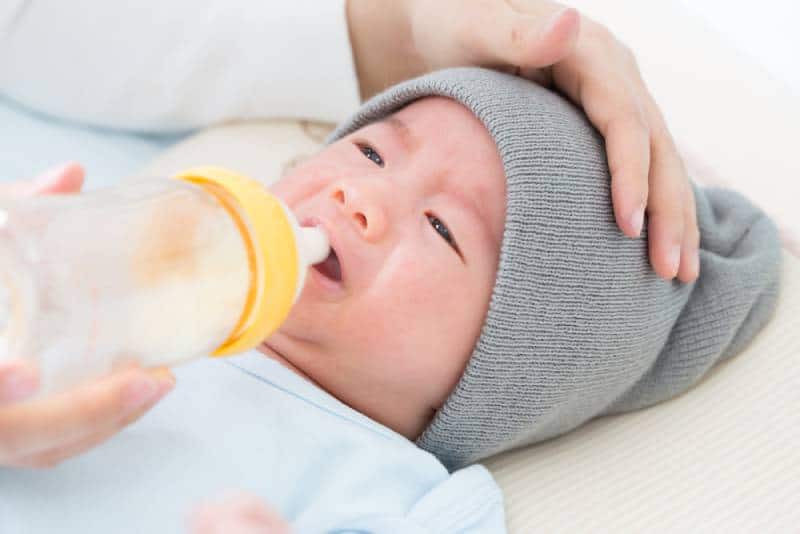 mother trying to give a milk bottle to her crying baby with hat