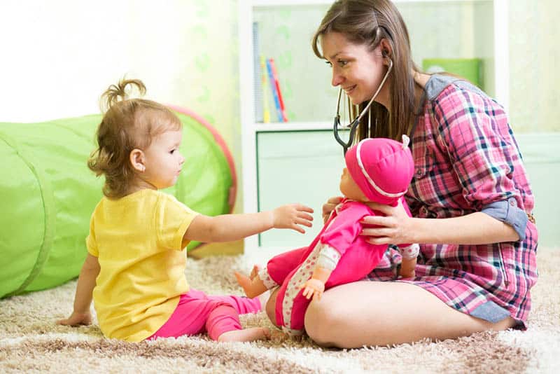 young mother playing a doctor with her baby girl on the floor with plastic baby toy