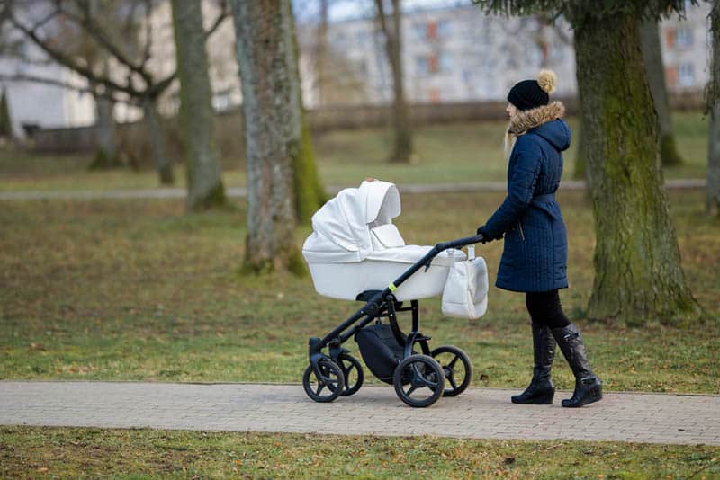 young mother pushing white baby stroller through the park