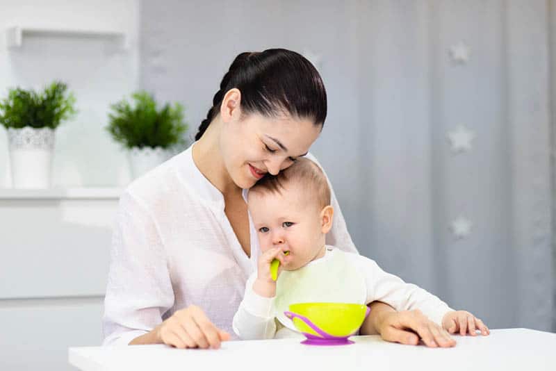 baby eating food with spoon while sitting in lap of happy mother