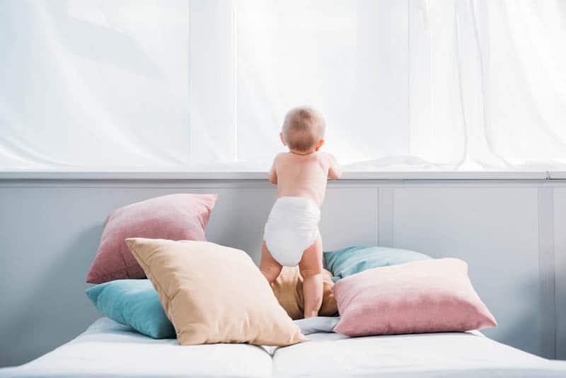 baby in diaper standing on the bed surrounded with colorful pillows